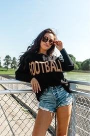 A woman in sunglasses and denim shorts leans on a metal fence, wearing the Panache Apparel Collection Black Football Sweater. The cozy crew neck sweater complements her casual style as she enjoys the sunny outdoor setting with trees and a field in the background.