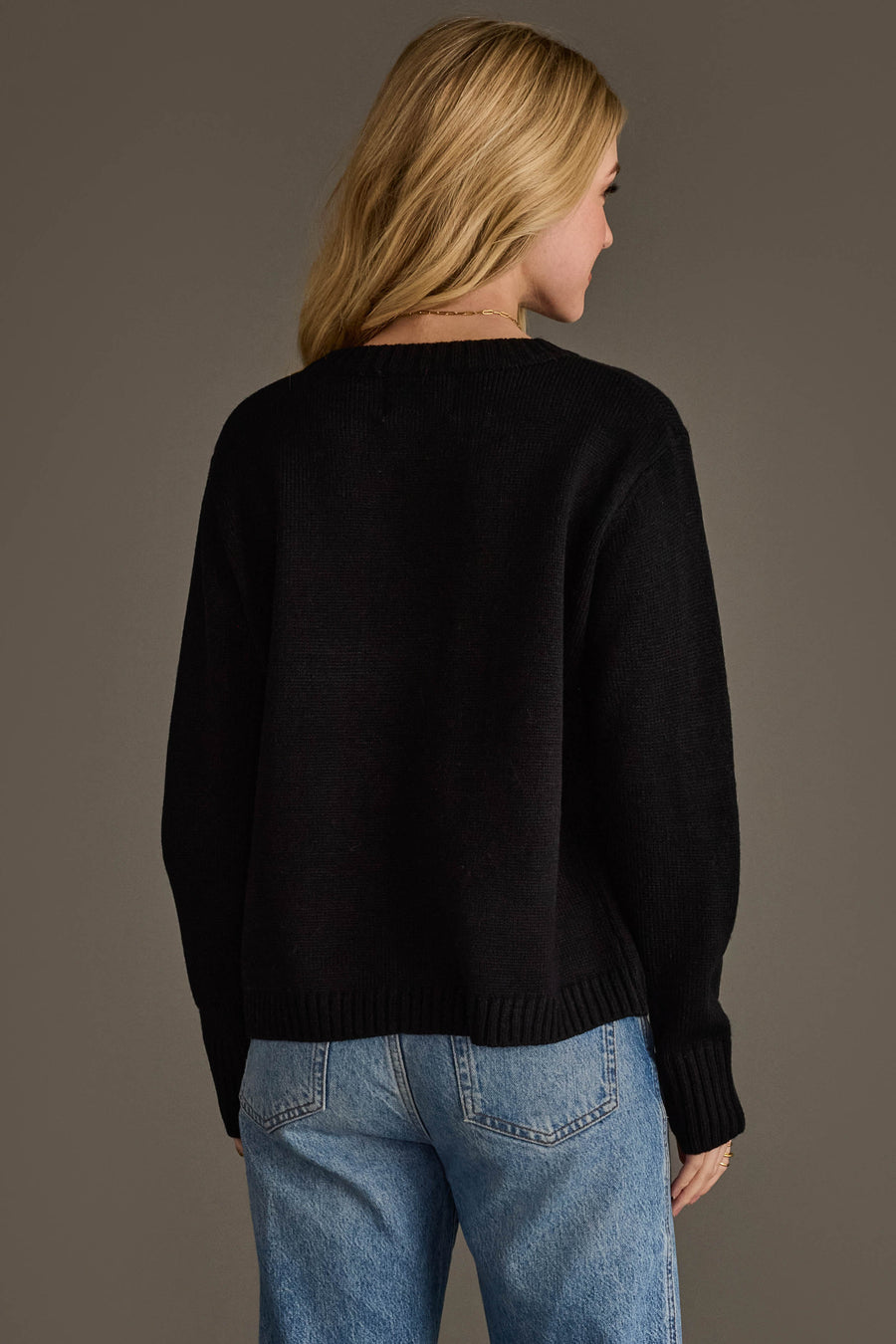 A woman with blonde hair, wearing the Panache Apparel Collection Black Football Sweater and light blue jeans, stands facing away from the camera against a plain, neutral background.