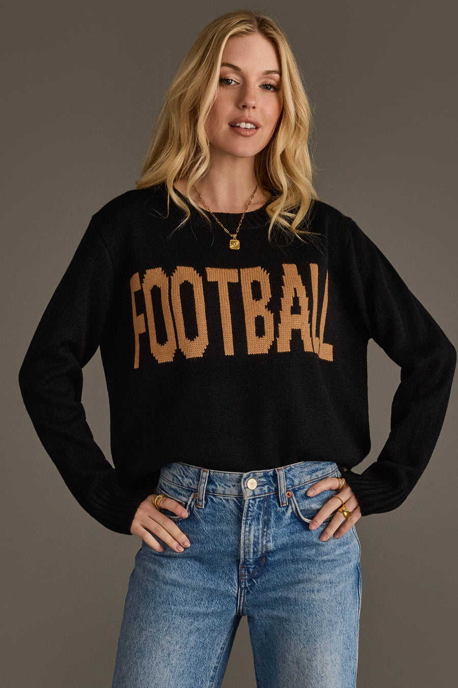 A woman with long blonde hair models the Panache Apparel Collection Black Football Sweater, featuring 'FOOTBALL' in tan letters, paired with light blue jeans as she poses with her hands on her hips against a plain background.