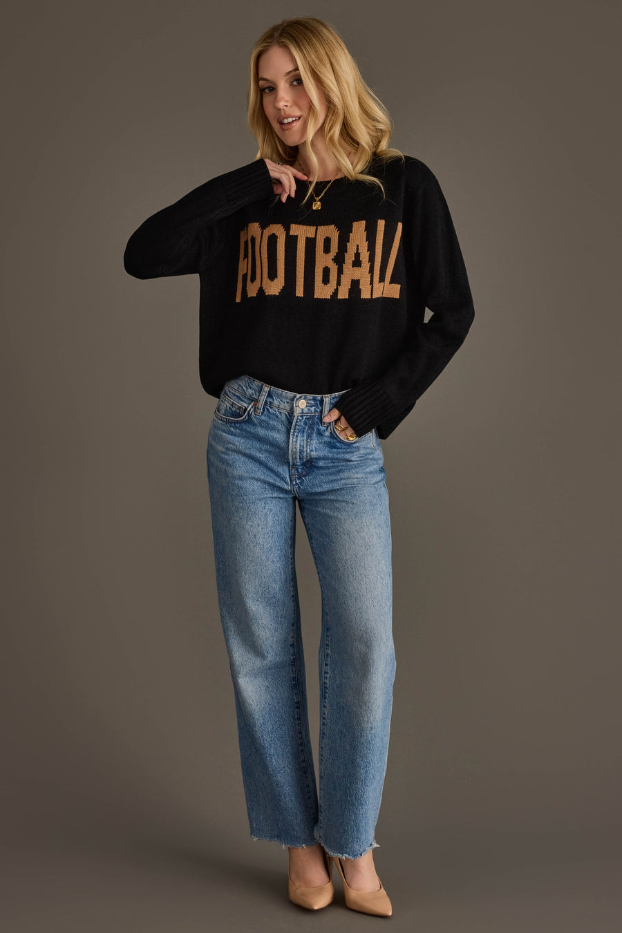 A woman with blonde hair, smiling and touching her necklace, stands against a gray background wearing the Panache Apparel Collection Black Football Sweater featuring "FOOTBALL" in tan letters, paired with blue jeans and beige heels.