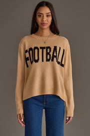 A woman models the Panache Apparel Collection's Tan Football Sweater, featuring the word "FOOTBALL" in bold black letters, paired with blue jeans and hoop earrings as she stands against a plain gray background.