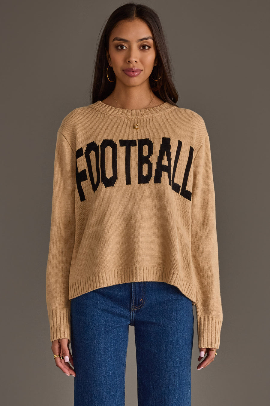 A woman models the Panache Apparel Collection's Tan Football Sweater, featuring the word "FOOTBALL" in bold black letters, paired with blue jeans and hoop earrings as she stands against a plain gray background.