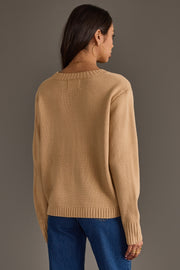 A woman with long dark hair, wearing large hoop earrings, a pair of blue jeans, and the Tan Football Sweater by Panache Apparel Collection, stands facing away from the camera against a plain gray background.