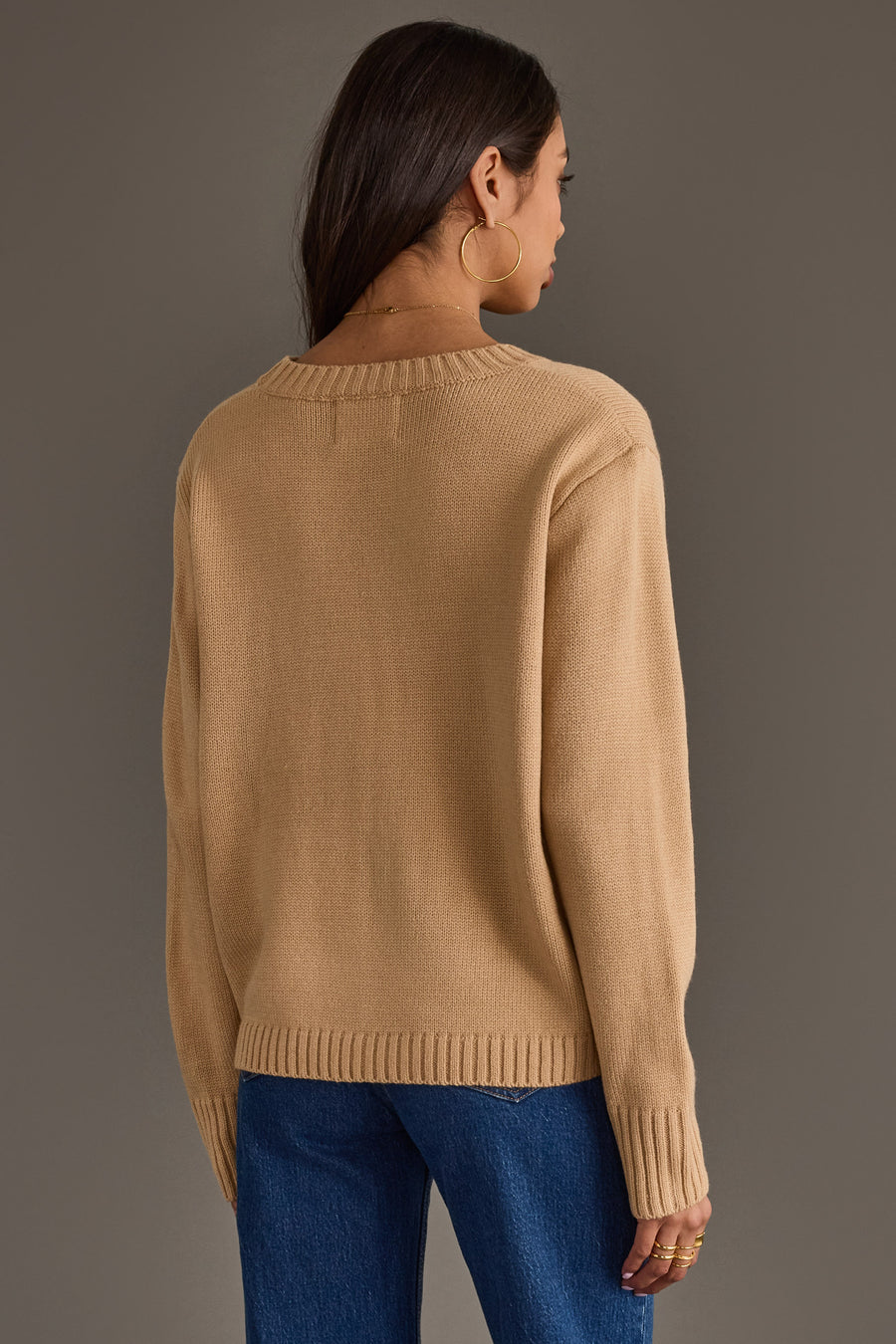 A woman with long dark hair, wearing large hoop earrings, a pair of blue jeans, and the Tan Football Sweater by Panache Apparel Collection, stands facing away from the camera against a plain gray background.