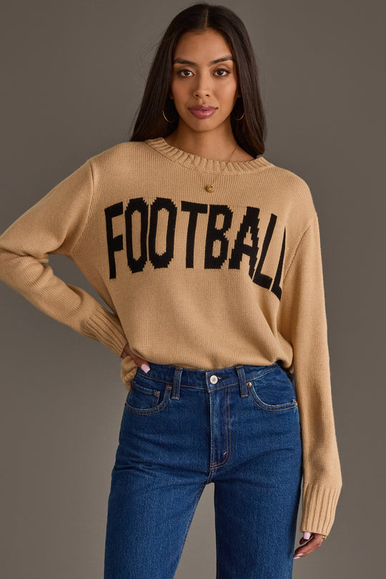 A woman models the Panache Apparel Collection Tan Football Sweater, featuring bold black "FOOTBALL" lettering, paired with blue jeans and posed against a plain gray background while looking at the camera.