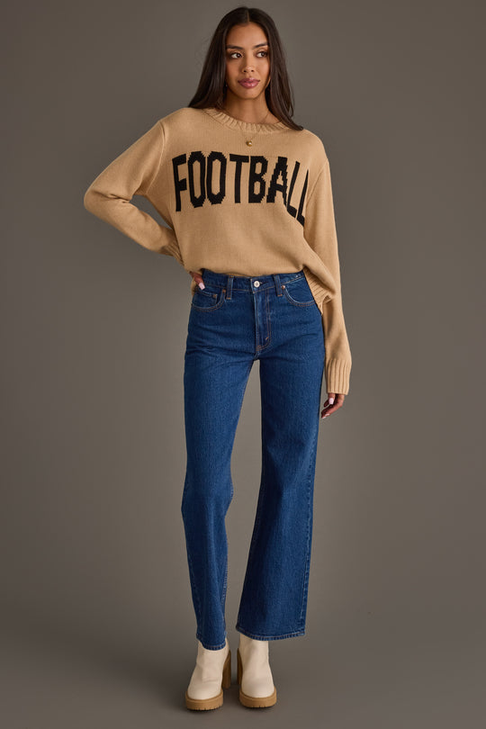 A woman stands against a gray background wearing the Panache Apparel Collection Tan Football Sweater, styled with blue high-waisted jeans and white ankle boots.