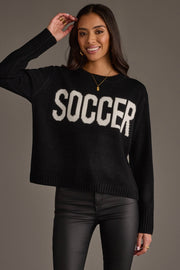 A smiling woman models the Panache Apparel Collection Soccer Sweater—a black top featuring "SOCCER" in bold white letters—paired with black pants against a plain background, her hand raised near her head.