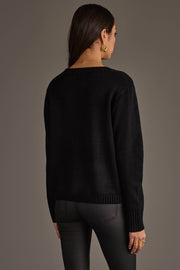 A woman with long brown hair, seen from behind, wears the Panache Apparel Collection Soccer Sweater paired with dark gray pants. She accessorizes with gold hoop earrings and rings, standing against a plain gray background.
