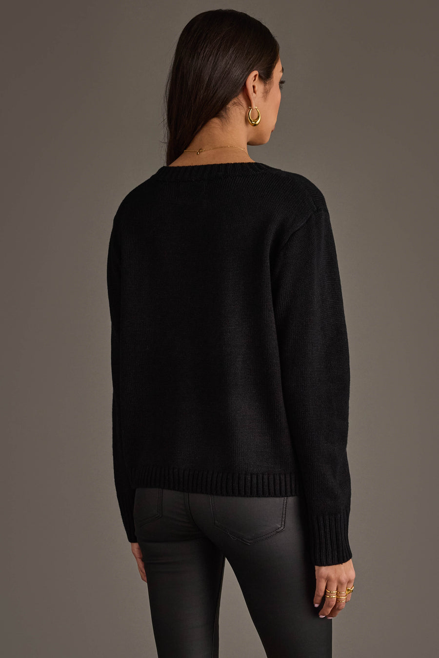 A woman with long brown hair, seen from behind, wears the Panache Apparel Collection Soccer Sweater paired with dark gray pants. She accessorizes with gold hoop earrings and rings, standing against a plain gray background.
