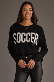 A woman with long dark hair is smiling and standing in front of a plain gray background, wearing the Panache Apparel Collection's Soccer Sweater—a black sweater featuring the word "SOCCER" in bold white letters—paired with black leather pants and gold jewelry.