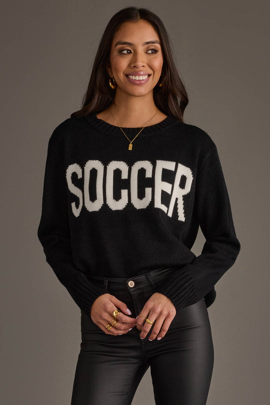 A woman with long dark hair is smiling and standing in front of a plain gray background, wearing the Panache Apparel Collection's Soccer Sweater—a black sweater featuring the word "SOCCER" in bold white letters—paired with black leather pants and gold jewelry.