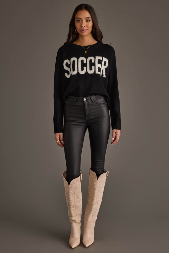 A woman with long dark hair stands against a gray background wearing the Panache Apparel Collection Soccer Sweater, styled with black leather pants and tall beige cowboy boots. She accessorizes with a necklace and rings.