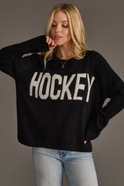 A woman models the Panache Apparel Collection Black Hockey Sweater, featuring bold white "HOCKEY" lettering, as she poses with one hand on her head and the other in her jeans pocket against a plain background.