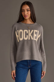 A woman with long dark hair wears the Panache Apparel Collection's Gray Hockey Sweater, featuring "HOCKEY" in bold cream letters, paired with blue jeans. She stands against a plain gray background, looking at the camera with a neutral expression.
