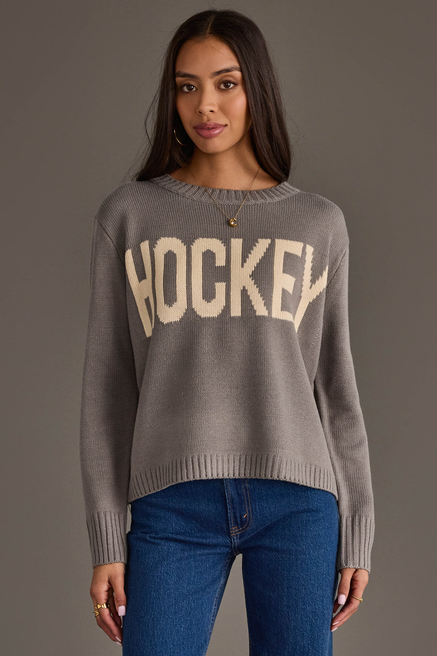A woman with long dark hair wears the Panache Apparel Collection's Gray Hockey Sweater, featuring "HOCKEY" in bold cream letters, paired with blue jeans. She stands against a plain gray background, looking at the camera with a neutral expression.