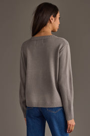 A woman with long dark hair, wearing large hoop earrings, blue jeans, and the Panache Apparel Collection Gray Hockey Sweater, stands facing away against a plain gray background.