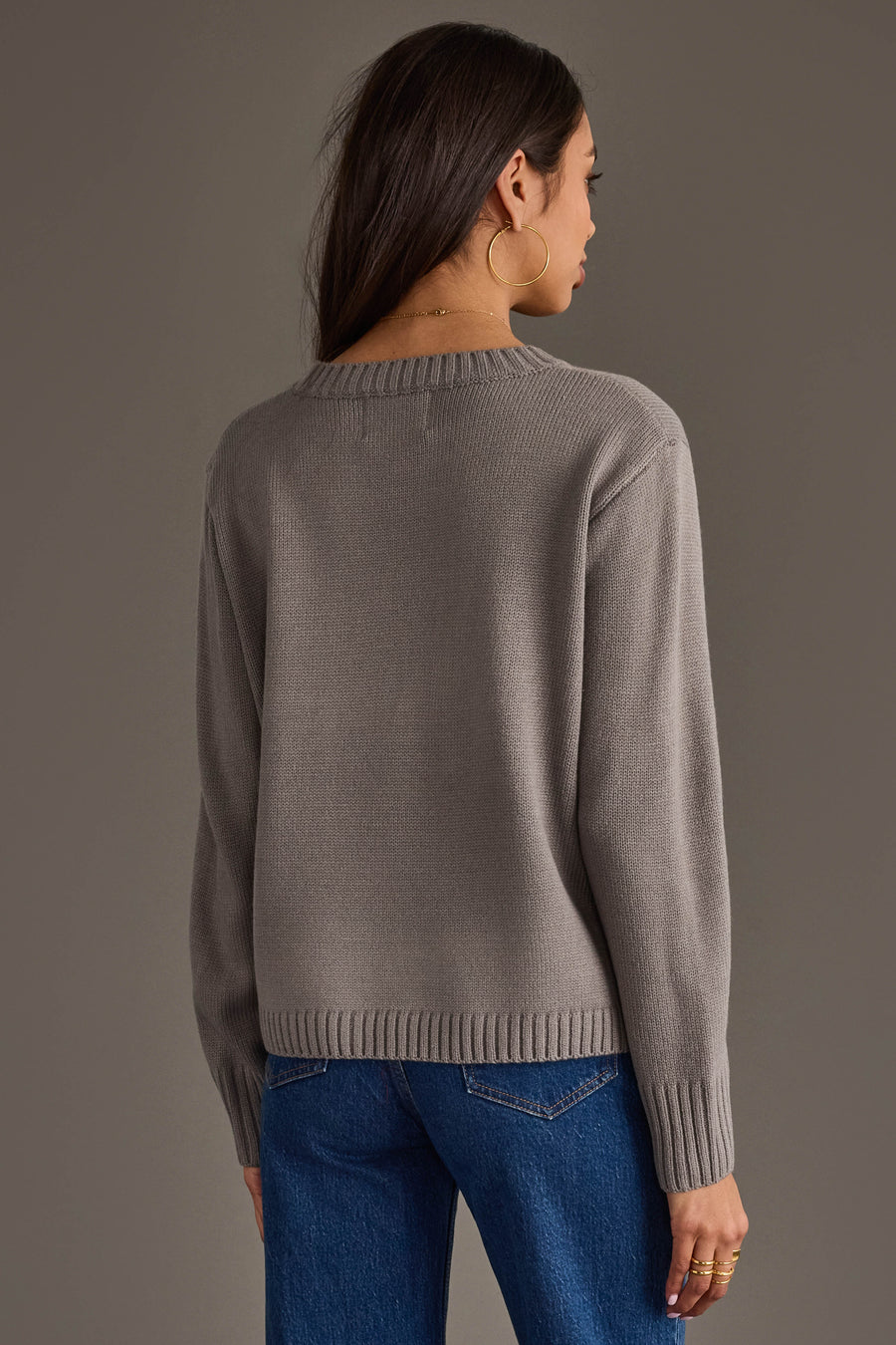 A woman with long dark hair, wearing large hoop earrings, blue jeans, and the Panache Apparel Collection Gray Hockey Sweater, stands facing away against a plain gray background.