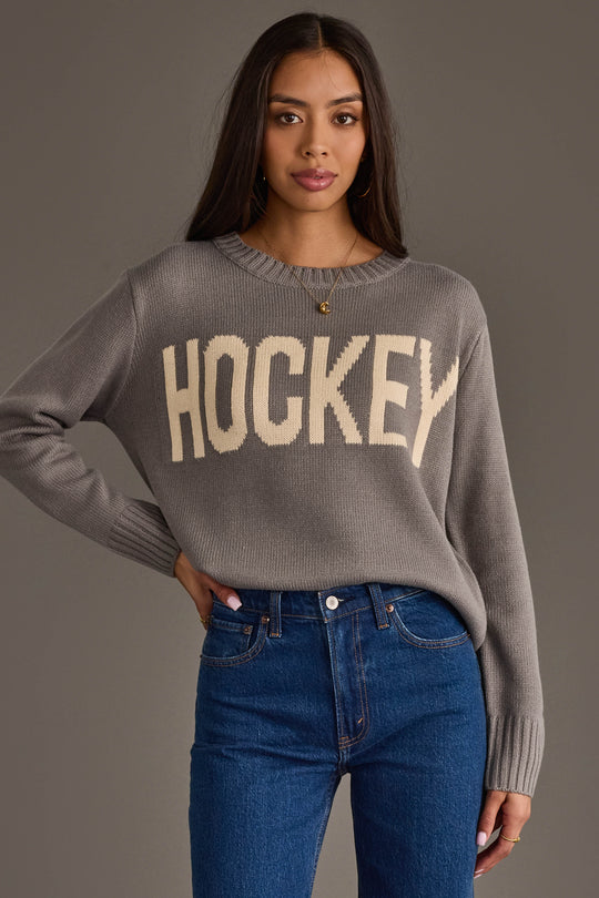 A woman with long dark hair models the Panache Apparel Collection Gray Hockey Sweater, featuring bold white "HOCKEY" lettering, paired with blue jeans and posed against a plain gray background.