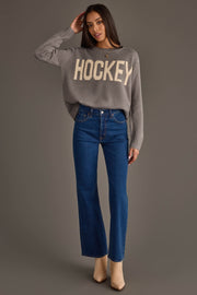 A woman stands against a gray background, wearing the Gray Hockey Sweater from Panache Apparel Collection, paired with blue high-waisted jeans and beige boots.