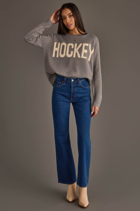 A woman stands against a gray background, wearing the Gray Hockey Sweater from Panache Apparel Collection, paired with blue high-waisted jeans and beige boots.