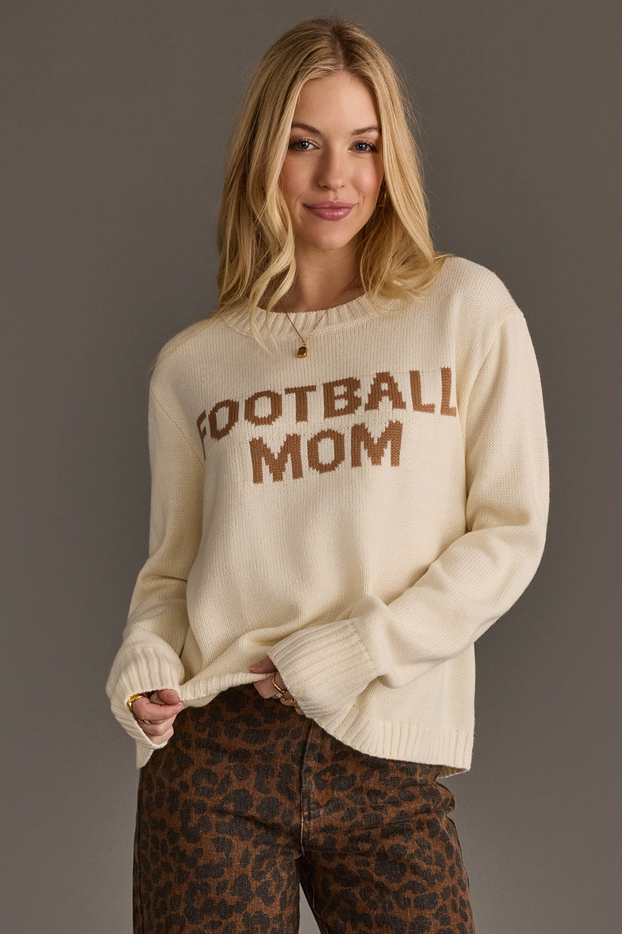 A woman with long blonde hair wears the Ivory Football Mom Sweater from Panache Apparel Collection along with brown leopard print pants, standing against a plain gray background.