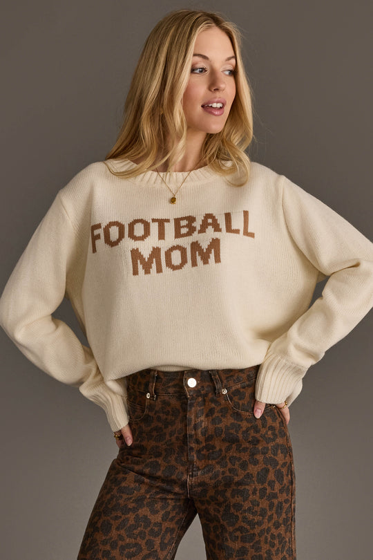 A woman is standing with her hands on her hips against a plain background, wearing the Panache Apparel Collection Ivory Football Mom Sweater—featuring "FOOTBALL MOM" in brown lettering—paired with high-waisted leopard print pants.