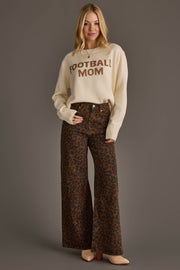 A woman with blonde hair stands smiling against a plain gray background, wearing the Panache Apparel Collection Ivory Football Mom Sweater paired with high-waisted, wide-leg brown leopard print pants and tan pointed-toe shoes.