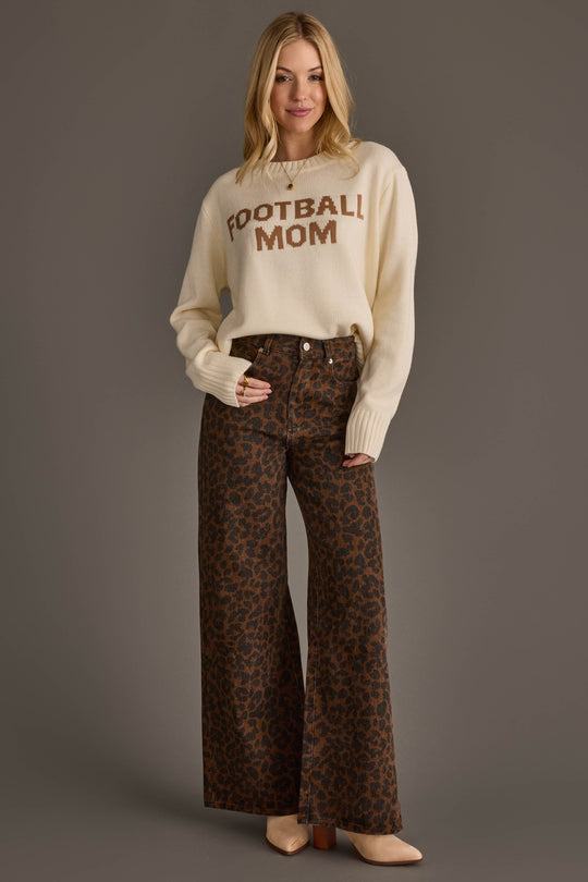 A woman with blonde hair stands smiling against a plain gray background, wearing the Panache Apparel Collection Ivory Football Mom Sweater paired with high-waisted, wide-leg brown leopard print pants and tan pointed-toe shoes.