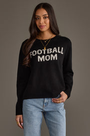 A woman models the Panache Apparel Collection Black & White Football Mom Sweater, featuring bold white lettering, as she stands against a plain background with one hand in her jeans pocket and a slight smile.