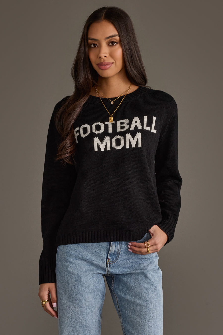 A woman models the Panache Apparel Collection Black & White Football Mom Sweater, featuring bold white lettering, as she stands against a plain background with one hand in her jeans pocket and a slight smile.