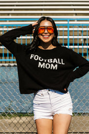 A smiling woman wearing sunglasses, the Black & White Football Mom Sweater by Panache Apparel Collection, and white shorts poses in front of stadium bleachers and a chain-link fence.