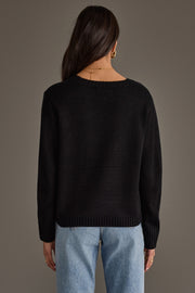A woman with long dark hair, wearing the Panache Apparel Collection Black & White Football Mom Sweater and light blue jeans, stands with her back to the camera against a plain gray background.