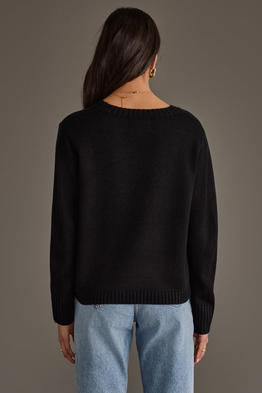 A woman with long dark hair, wearing the Panache Apparel Collection Black & White Football Mom Sweater and light blue jeans, stands with her back to the camera against a plain gray background.