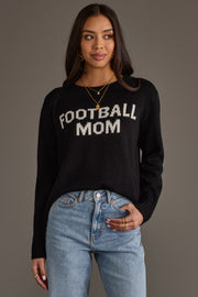 A woman models the Panache Apparel Collection Black & White Football Mom Sweater, paired with blue jeans and gold jewelry, her hair worn loose as she stands against a plain gray background.