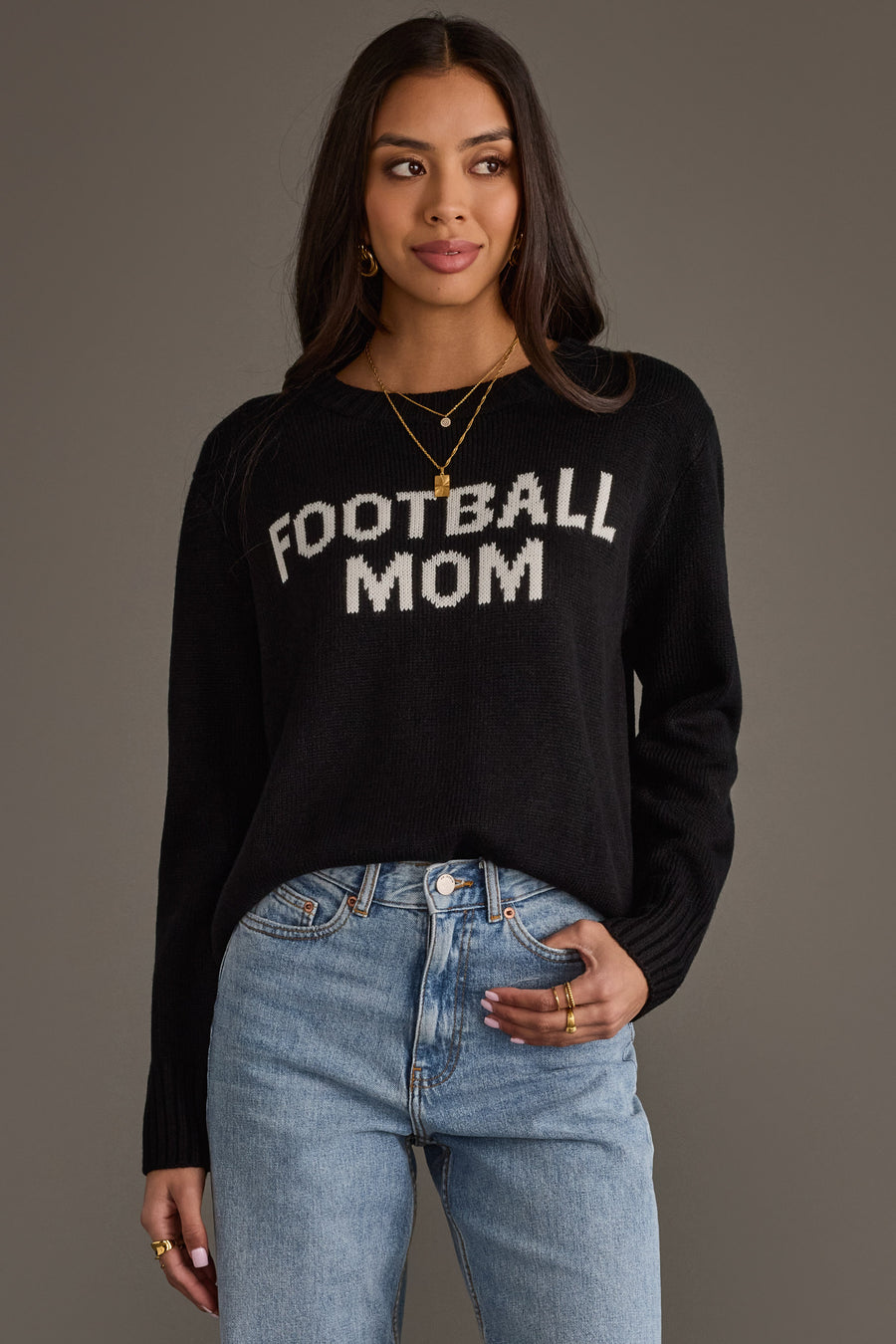 A woman models the Panache Apparel Collection Black & White Football Mom Sweater, paired with blue jeans and gold jewelry, her hair worn loose as she stands against a plain gray background.