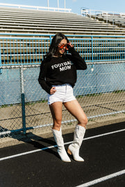 A woman wearing sunglasses, a pair of white shorts, and white boots stands on a track near the bleachers, posing with one hand on her sunglasses. She is dressed in the Panache Apparel Collection Black & White Football Mom Sweater.