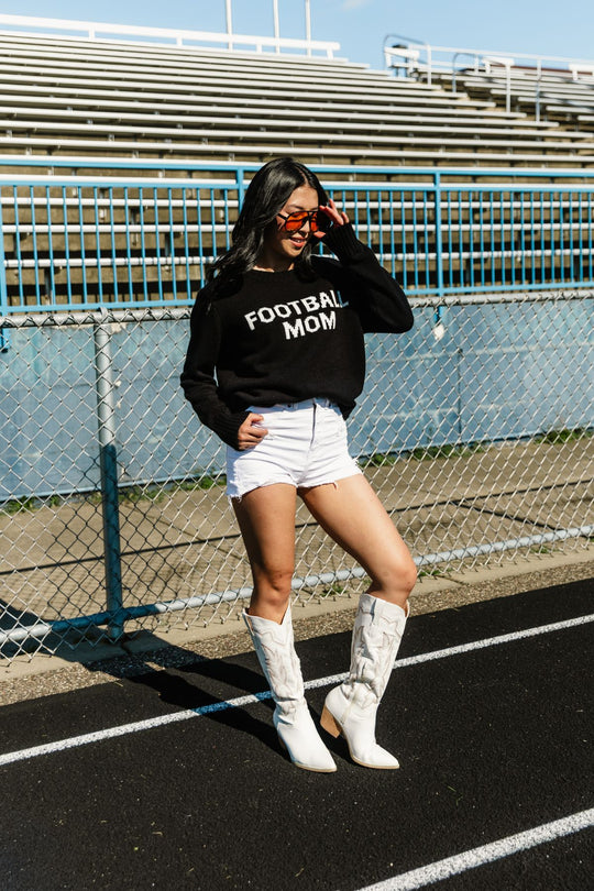 A woman wearing sunglasses, a pair of white shorts, and white boots stands on a track near the bleachers, posing with one hand on her sunglasses. She is dressed in the Panache Apparel Collection Black & White Football Mom Sweater.