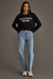 A woman stands against a gray background wearing the Panache Apparel Collection Black & White Football Mom Sweater, paired with light blue jeans, white sneakers, and a gold necklace, her hands tucked into her pockets.