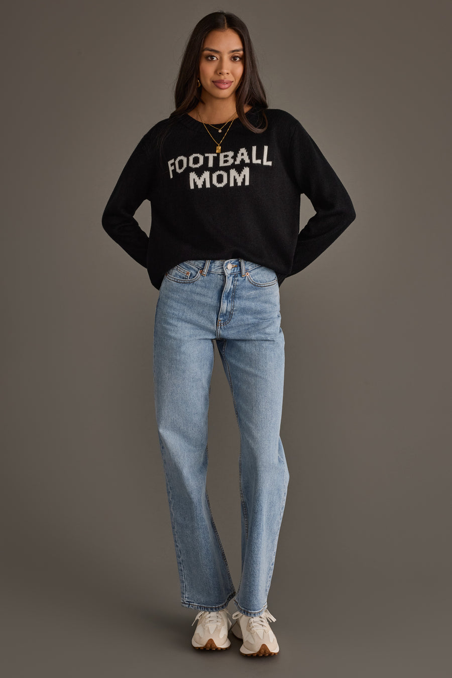 A woman stands against a gray background wearing the Panache Apparel Collection Black & White Football Mom Sweater, paired with light blue jeans, white sneakers, and a gold necklace, her hands tucked into her pockets.