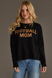 A woman with long blonde hair is wearing the Panache Apparel Collection's Black & Tan Football Mom Sweater, paired with blue jeans and gold jewelry. She is smiling while standing against a plain gray background.