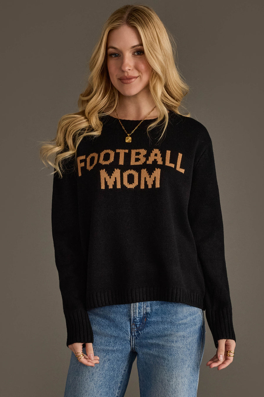 A woman with long blonde hair is wearing the Panache Apparel Collection's Black & Tan Football Mom Sweater, paired with blue jeans and gold jewelry. She is smiling while standing against a plain gray background.