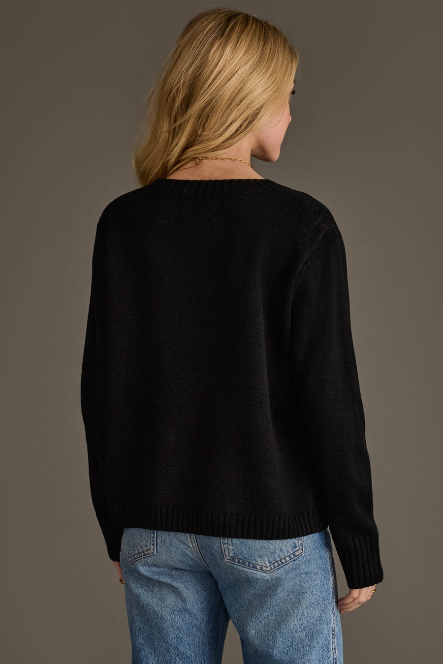 A person with long blonde hair, wearing the Black & Tan Football Mom Sweater from Panache Apparel Collection and blue jeans, stands facing away from the camera against a plain background.