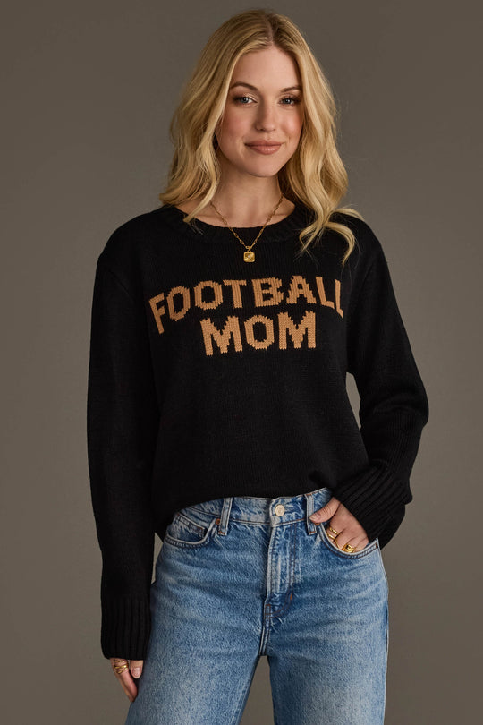 A woman with long blonde hair is standing against a plain background, smiling with one hand in her pocket. She is wearing blue jeans and the Panache Apparel Collection Black & Tan Football Mom Sweater.