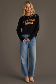 A woman with long blonde hair is smiling against a plain gray background, wearing the Panache Apparel Collection Black & Tan Football Mom Sweater, blue jeans, and beige heels.