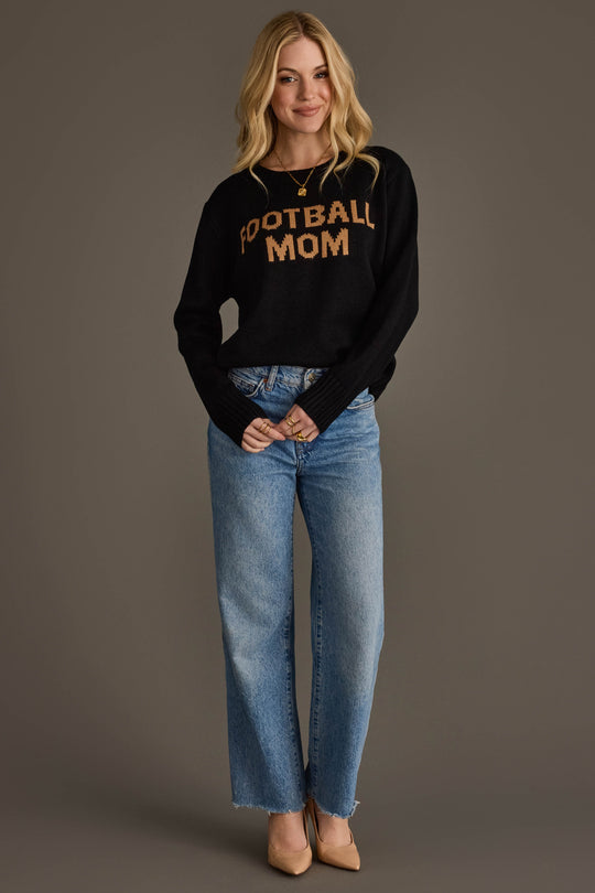 A woman with long blonde hair is smiling against a plain gray background, wearing the Panache Apparel Collection Black & Tan Football Mom Sweater, blue jeans, and beige heels.