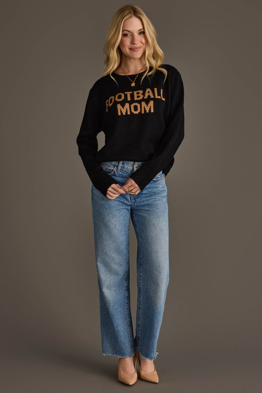 A woman with long blonde hair is smiling against a plain gray background, wearing the Panache Apparel Collection Black & Tan Football Mom Sweater, blue jeans, and beige heels.