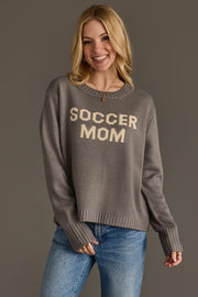 A smiling woman with blonde hair is wearing the Panache Apparel Collection "Soccer Mom" sweater in gray, featuring beige lettering, paired with blue jeans and standing against a plain dark background.