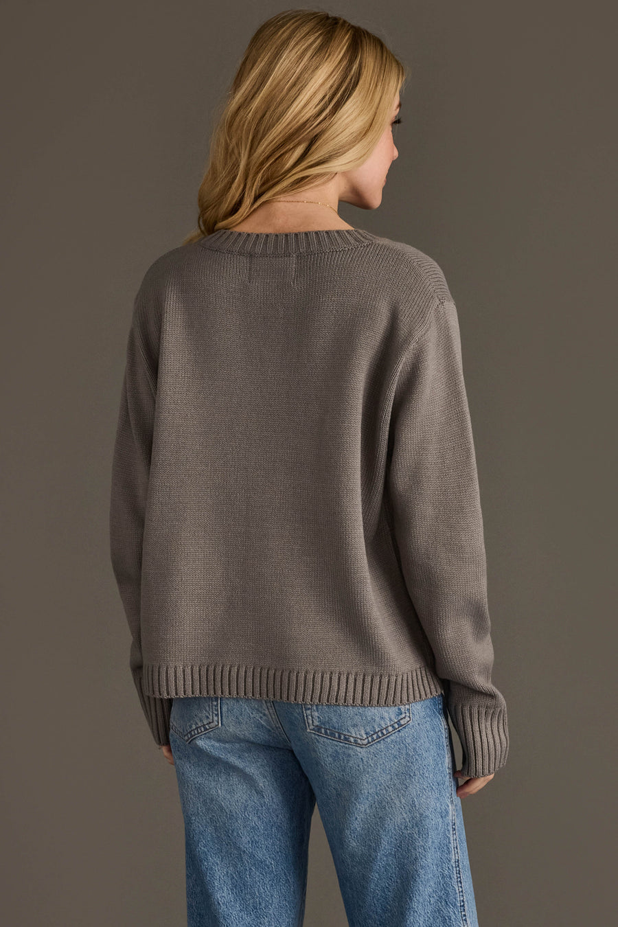 A person with long blonde hair, wearing the Panache Apparel Collection's Soccer Mom Sweater in gray and blue jeans, stands facing away from the camera against a plain background.