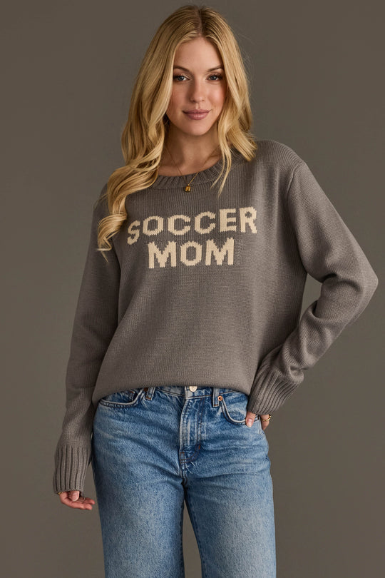 A woman with long blonde hair models the Panache Apparel Collection's Soccer Mom Sweater—a gray pullover featuring "SOCCER MOM" in white letters—paired with blue jeans, set against a plain gray background.