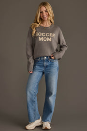 A smiling woman with long blonde hair models the Panache Apparel Collection Soccer Mom Sweater in gray, paired with blue jeans and sneakers, standing against a plain brown background.